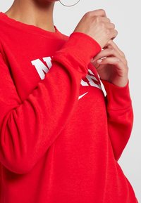 Sudadera roja de tejido suave, con texto y logo "NIKE" en blanco, puños acanalados y un corte holgado.