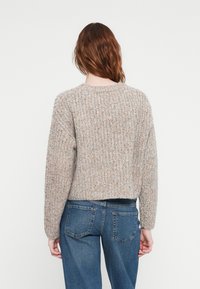 Gai pelēks tekstūrēts džemperis ar apaļu kakla izgriezumu un garām piedurknēm, ar smalkiem krāsainiem flekiem, kombinēts ar zilām džinsu biksēm.