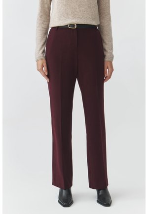 Persoon draagt hooggetailleerde rechtse bordeauxrode broek met zwarte riem, beige trui met lange mouwen en zwarte laarzen met puntige neus tegen een witte achtergrond.