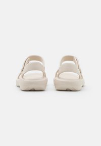 Lys beige sandaler med åpen bakdel, justerbar stropp, teksturert overdelsmateriale og polstret såle synlig fra bakvinkelen.