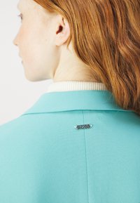 Blazer turchese con colletto a coste bianco a contrasto. Presenta una spilla con il logo "BOSS" in argento sul retro, evidenziando il brand del capo.
