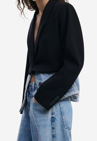 Schwarzer, kurz geschnittener Blazer mit Schalkragen, kombiniert mit einer hellblauen Denimjacke. Der Stoff wirkt glatt mit einer strukturierten Passform und sichtbaren Knöpfen.