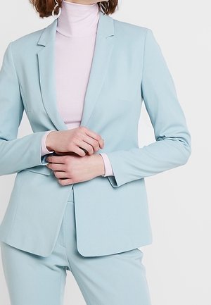 Blazer bleu clair avec une coupe ajustée, présentant une fermeture à un bouton, des revers notched et une texture lisse. Porté par-dessus un col roulé rose.