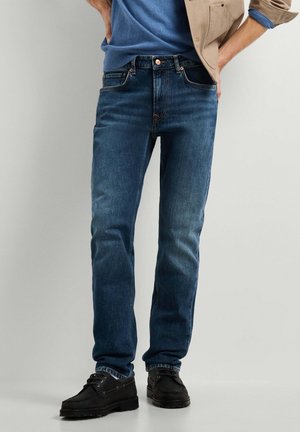 Mann trägt blaue Jeans, schwarze Schnürschuhe, blaues Hemd und beige Jacke mit den Händen in den Taschen vor schlichtem Hintergrund.