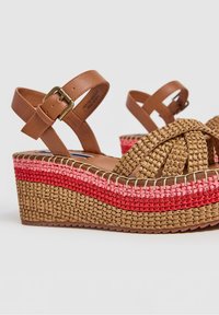 Sandales compensées marron tressées avec des rayures tressées roses et rouges sur la semelle et une bride de cheville en cuir avec une boucle.