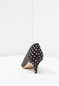 Black kitten heel shoe with white polka dots, pointed toe, and short slender heel on white surface against white background.