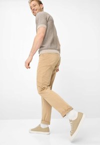 Beige katoenen broek met opgerolde pijpen, gecombineerd met beige sneakers en witte sokken. De bruine gebreide korte mouw trui heeft kleurrijke patronen.