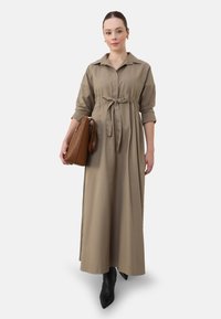 Langsleeve, beige jurk met een overhemdkraag en een ceintuur in de taille. Model houdt een brown tas vast en draagt spitse zwarte enkellaarsjes. Gladde textuur.