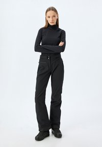 Zwarte top met lange mouwen en hoge halslijn, gecombineerd met zwarte, losse broek met een tailleband en ritszakken. Zwarte schoenen.