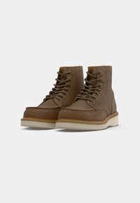 Un paio di stivali alla caviglia in suede marrone, con lacci e suole in gomma beige chiaro, su uno sfondo bianco, posizionati affiancati.