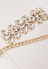 Kopertówka ozdobiona dużymi przezroczystymi kryształami w różnych kształtach, złotym metalowym wykończeniem i łańcuszkiem na tle miękkiej beżowej tkaniny.