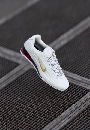 Basket Nike blanche avec le swoosh doré et des lacets blancs flottant au-dessus d’un fond texturé en grille métallique sombre.