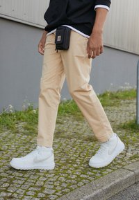 Beige cotton pants and white high-top Nike sneakers. A black pouch hangs from the waist, worn over a dark hoodie with a white inner layer.
