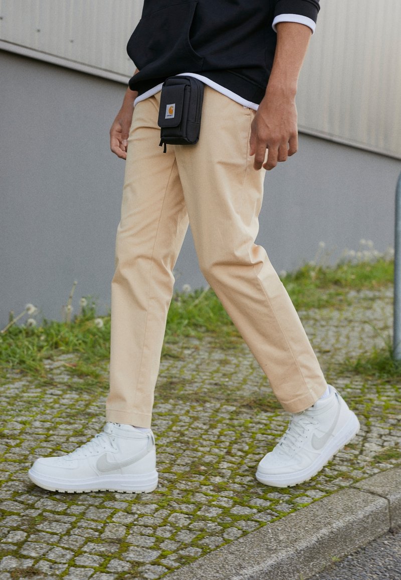 Beige cotton pants and white high-top Nike sneakers. A black pouch hangs from the waist, worn over a dark hoodie with a white inner layer.