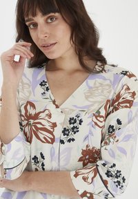Blusa floreale con scollo a V, maniche a tre quarti, di colore crema con grandi fiori bordeaux e blu. Tessuto morbido con una texture leggermente lucida.