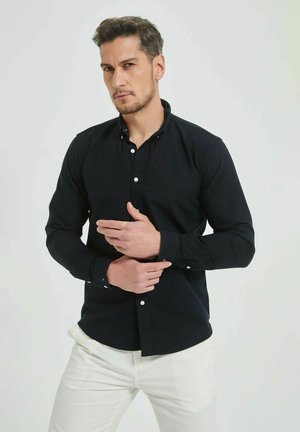 Chemise noire à boutons avec manches longues et une texture subtile, ornée de boutons blancs, associée à un pantalon de couleur claire.