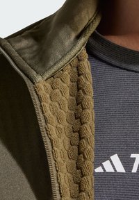 Collo di giacca verde oliva con foderatura beige a motivo a nido d'ape. Camicia grigia sotto, con logo bianco e scollo a coste.