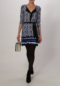 Sukienka z geometrycznym czarno-białym wzorem, dekoltem w kształcie litery V, rękawami o długości trzech kwart, zaakcentowaną talią, wykończona niebieskim paskiem na dole.