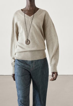 Person wearing a beige V-neck sweater, blue jeans, and a long necklace with a round wooden pendant, standing against a plain background.