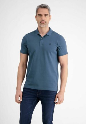 Mann mit kurzem grauem Haar und Bart, der ein blaues Poloshirt und dunkle Jeans trägt, vor einem weißen Hintergrund stehend.