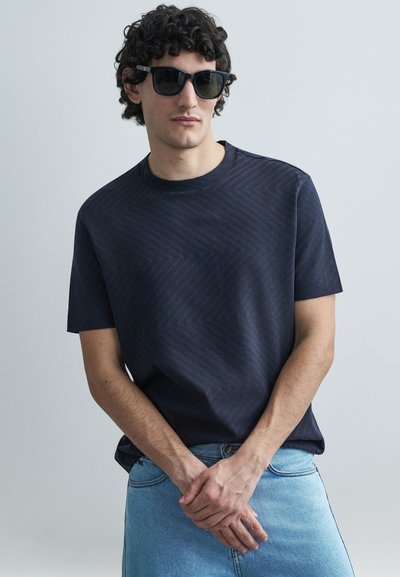 Granatowy t-shirt z krótkim rękawem o fakturze zygzakowatego wzoru, w połączeniu z jasnoniebieską spódnicą dżinsową. Modelka nosi czarne okulary przeciwsłoneczne.