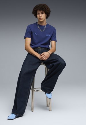 Giovane uomo con capelli ricci seduto su uno sgabello, indossa una t-shirt blu Puma, jeans larghi scuri, scarpe azzurro chiaro e catene d'argento.