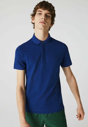 Camisa polo azul con cuello clásico y mangas cortas. Presenta un logo bordado sutil y una tela lisa y texturada.