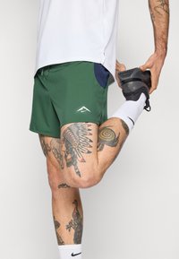 Pantalones cortos deportivos verdes con un bolsillo azul, que presentan un logo blanco. El usuario estira una pierna mientras lleva puesta una zapatilla negra y calcetines blancos.