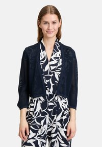 Marineblauer Strickcardigan mit Dreiviertelärmeln, offenem Design und zarter Netzstruktur über einer floral gemusterte weißen Bluse.