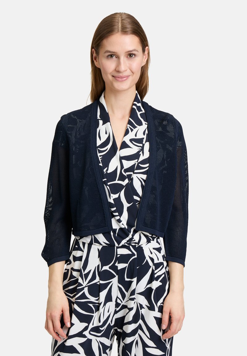 Marineblauer Strickcardigan mit Dreiviertelärmeln, offenem Design und zarter Netzstruktur über einer floral gemusterte weißen Bluse.