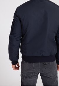 Muž v tmavě modré bomberce a vybledlých černých džínách, zobrazený zezadu na jednobarevném světlém pozadí.