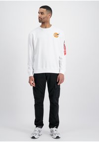 Biały sweatshirt z okrągłym dekoltem z logo na piersi, zestawiony z czarnymi spodniami cargo i białymi sportowymi butami. Widoczny czerwony zamek.