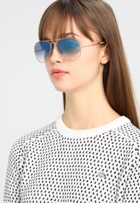 Ochelari de soare aviator cu ramă aurie și lentile cu gradient albastru, purtați de un model într-un pulover alb cu modele de puncte negre și un logo.