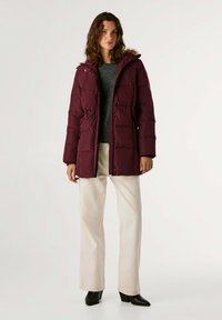 Chaqueta de invierno acolchada burdeos con capucha de piel sintética, cierre frontal con cremallera y bolsillos laterales, llevada sobre un suéter gris y pantalones crema.