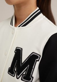 Veste varsity blanche avec manches noires, arborant un écusson "M" texturé en noir. Col côtelé noir et blanc rayé avec fermeture à boutons-pression.