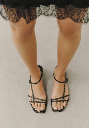 Jambes de femme portant une jupe en dentelle noire et des sandales noires à brides, debout sur un sol gris clair.