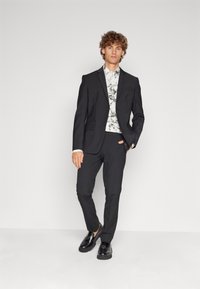 Schwarzer, taillierter Anzug mit einreihigem Jacket, dezentem Taschenstil, kombiniert mit einem weiß-schwarz marmorierten Hemd und schwarzen Schuhen.