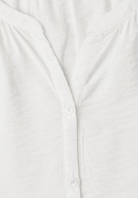 Blouse blanche à col en V avec une petite patte de boutonnage et des détails subtils de tissu froncé près de l'encolure.