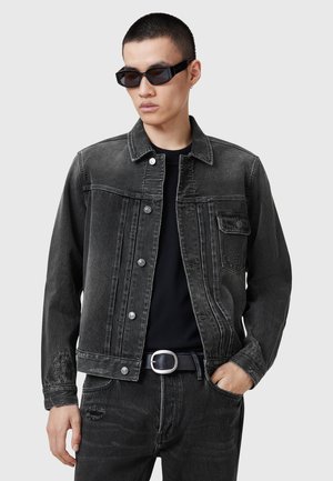 Giacca di denim nera con colletto classico, tasca frontale e dettagli con bottoni. Indossata sopra una camicia nera dal taglio slim.