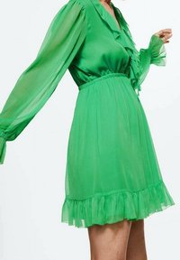 Frau in einem knielangen, langärmeligen grünen Kleid mit rüschigem Kragen und Saum, transparenten Ärmeln und tailliertem Bund.