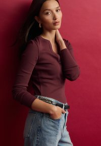 Bordo matinė ilgomis rankovėmis palaidinė su mygtukų detale ir apvaliu iškirpimu, derinama su šviesiai mėlynomis nusidėvėjusiomis džinsomis, pabrėžiama juodu diržu.