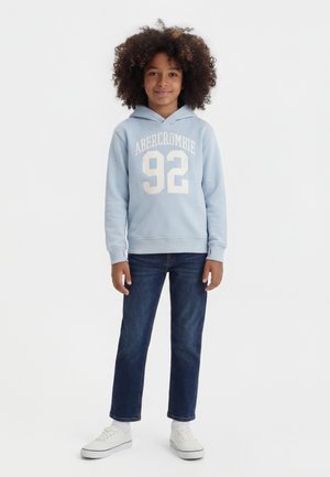 Παιδί με σγουρά μαλλιά που φοράει γαλάζιο hoodie Abercrombie, σκούρα μπλε τζιν και λευκά αθλητικά παπούτσια, στέκεται μπροστά σε απλό φόντο.