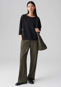 Schwarzes langärmliges Oberteil, olivgrüne weit geschnittene Hose mit gerippter Textur und eine strukturiert gestaltete dunkle Handtasche. Das Model trägt schwarze Slipper.
