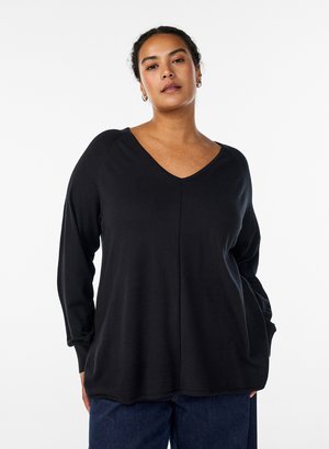 Kvinde med mørkt hår trukket tilbage, iført guldfarvede øreringe i ring, en sort V-hals langærmet top og mørkeblå jeans, stående mod en hvid baggrund.