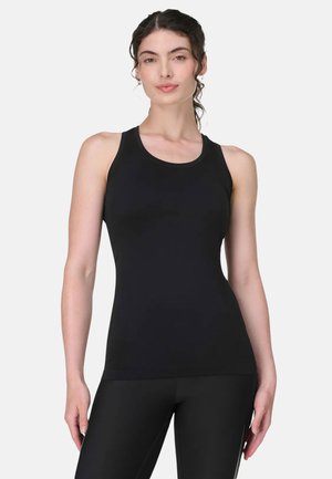 Vrouw draagt een strak passend zwart mouwloos tanktopje en zwarte legging, staat met ontspannen armen tegen een effen lichte achtergrond.