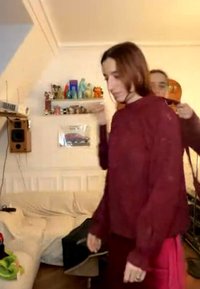 Une personne joue du violon derrière une autre personne portant un pull bordeaux dans une pièce avec des étagères contenant des figurines colorées.