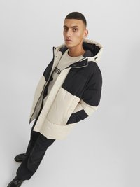 Pufferjack in crème en zwart, met een afneembare capuchon, ritssluiting en grote voorkanten. Matte textuur met een gestikt patroon.