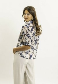 Blouse transparente à motifs floraux en crème avec des accents bleu et marron ; présente un col et des manches retroussées, associée à un pantalon clair.