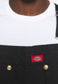 Salopette nera con accessori dorati, dotata di una patch logo rossa Dickies. Sotto, è visibile una maglietta bianca semplice.