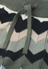 Gros plan d’un tissu tricoté avec des motifs chevrons verts, noirs, beiges et blancs, avec un cordon vert noué.
